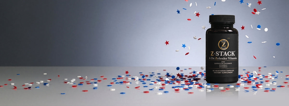 A black bottle of Z-Stack Ultra Advanced Vitamin with a black cap, standing on a reflective surface surrounded by scattered red, white, and blue confetti against a gray gradient background.