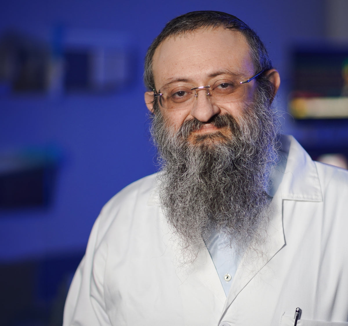 A portrait of a man with a long gray beard and glasses, wearing a white lab coat over a light blue shirt, set against a dark blue background.